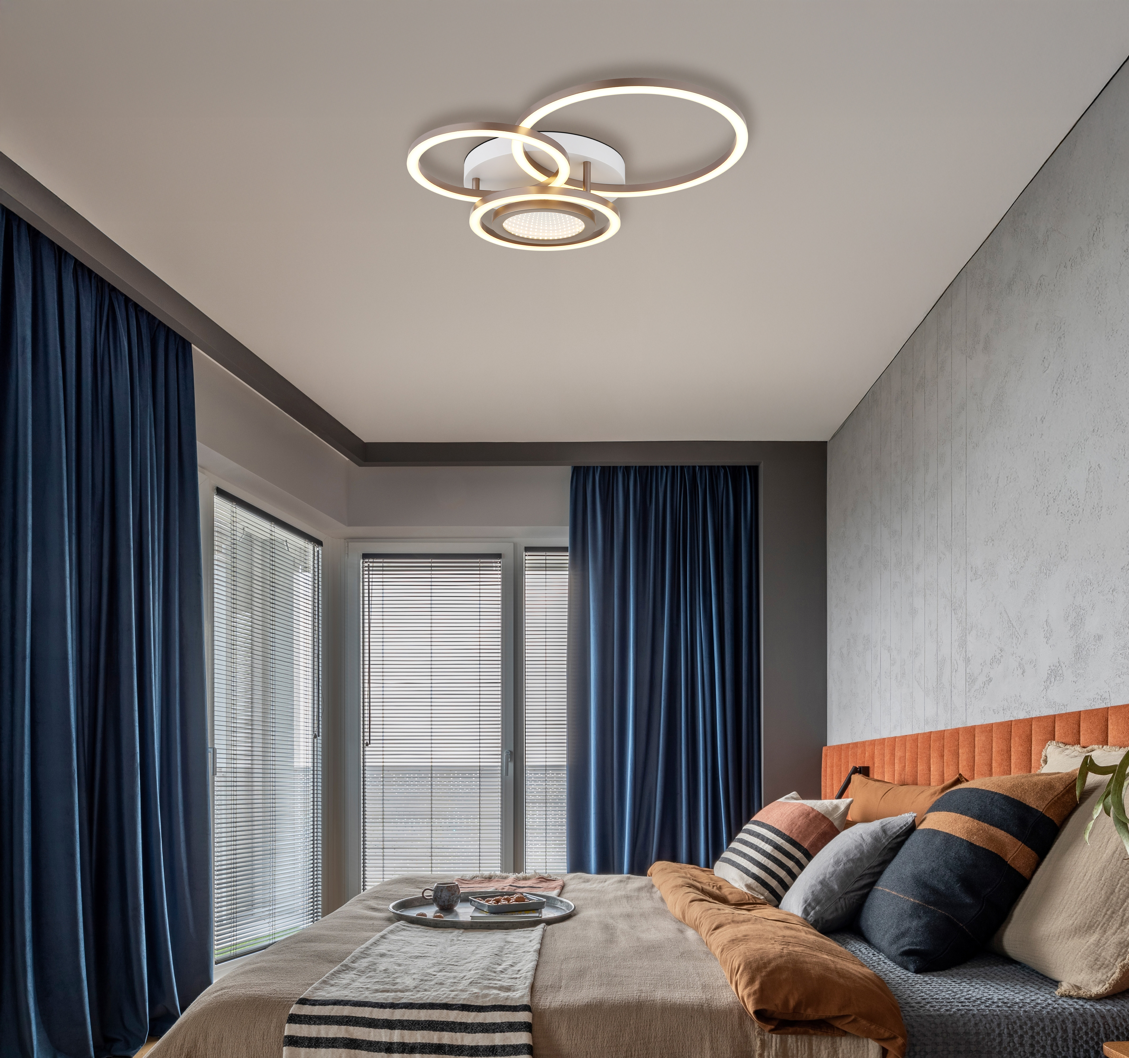 Schlafzimmer mit blauen Vorhängen, großen Fenstern, beigem Bett und runder Deckenleuchte aus Leuchtkreisen in warmweiß.